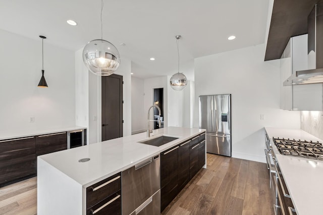 kitchen with sink, wall chimney range hood, appliances with stainless steel finishes, an island with sink, and decorative light fixtures