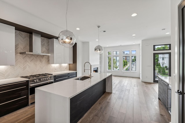 kitchen with wall chimney exhaust hood, stainless steel gas range, sink, decorative light fixtures, and a kitchen island with sink