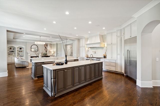 kitchen featuring custom exhaust hood, hanging light fixtures, an island with sink, dark wood-type flooring, and built in fridge