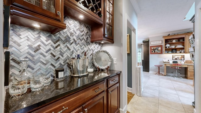 bar with built in desk, tasteful backsplash, dark stone countertops, light tile patterned floors, and crown molding