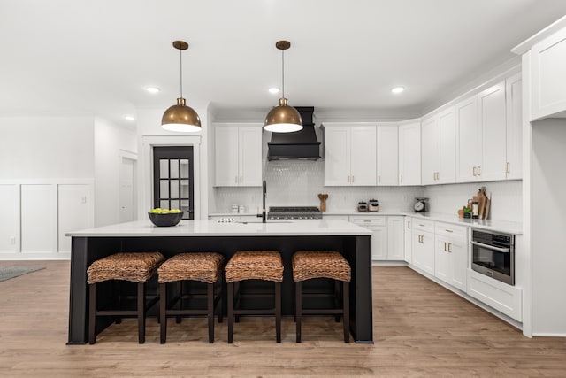 kitchen featuring stainless steel oven, premium range hood, an island with sink, and white cabinetry