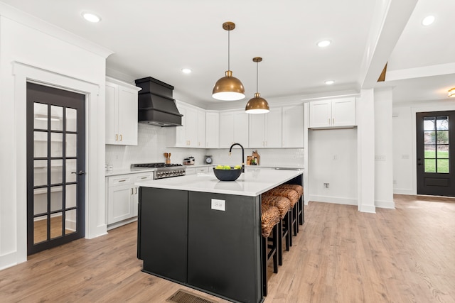 kitchen featuring white cabinetry, custom range hood, an island with sink, and hanging light fixtures