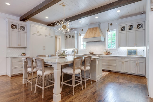 kitchen featuring premium range hood, beamed ceiling, white cabinetry, hanging light fixtures, and a center island with sink