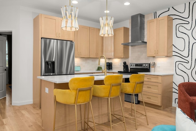 kitchen with appliances with stainless steel finishes, light brown cabinetry, a kitchen bar, hanging light fixtures, and wall chimney exhaust hood