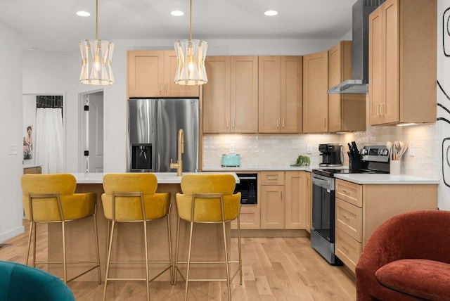 kitchen with wall chimney exhaust hood, decorative light fixtures, light brown cabinets, appliances with stainless steel finishes, and light hardwood / wood-style floors