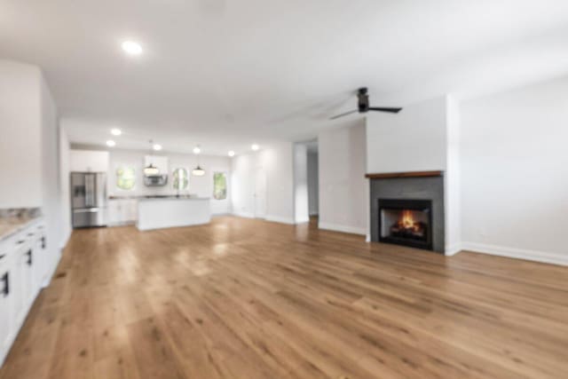 unfurnished living room with recessed lighting, a warm lit fireplace, light wood-style flooring, baseboards, and a ceiling fan