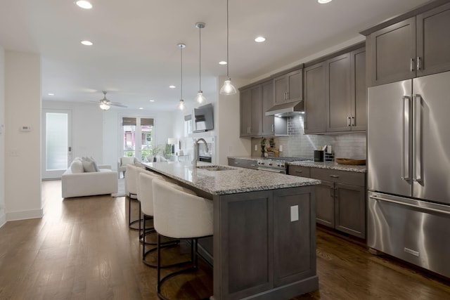 kitchen with sink, high end appliances, light stone counters, hanging light fixtures, and an island with sink