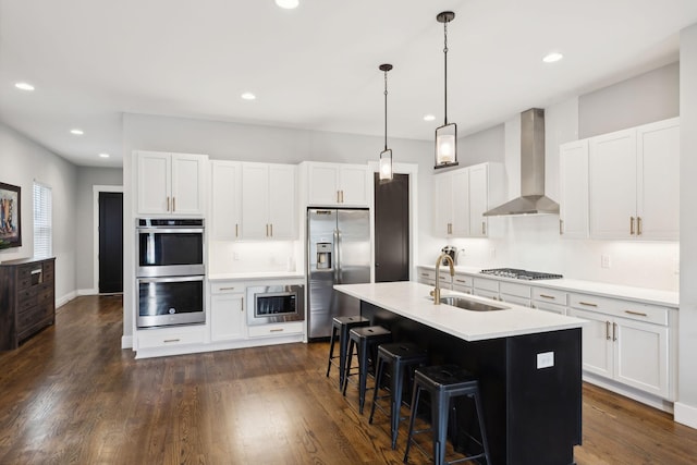 kitchen featuring appliances with stainless steel finishes, white cabinetry, sink, wall chimney range hood, and a center island with sink