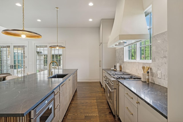 kitchen with pendant lighting, high end stainless steel range oven, dark stone countertops, an island with sink, and custom range hood