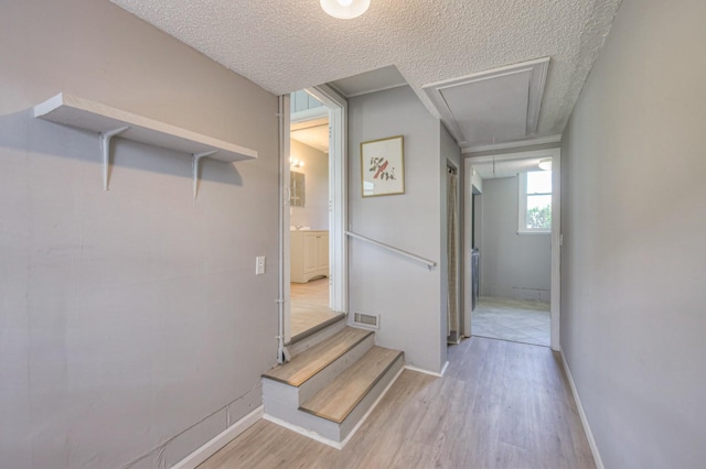 hallway featuring light wood finished floors, attic access, visible vents, baseboards, and a textured ceiling