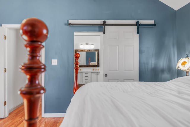 bedroom with a barn door, baseboards, vaulted ceiling, light wood finished floors, and ensuite bath