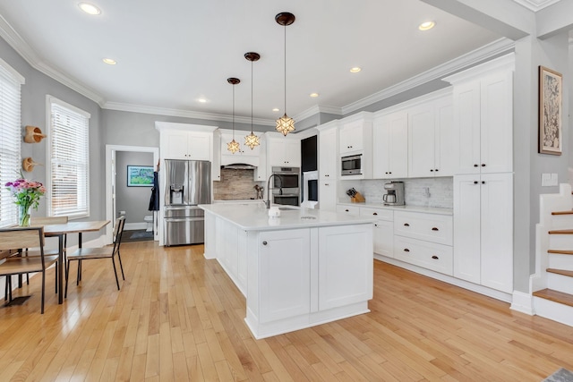 kitchen featuring appliances with stainless steel finishes, an island with sink, white cabinets, hanging light fixtures, and light hardwood / wood-style flooring