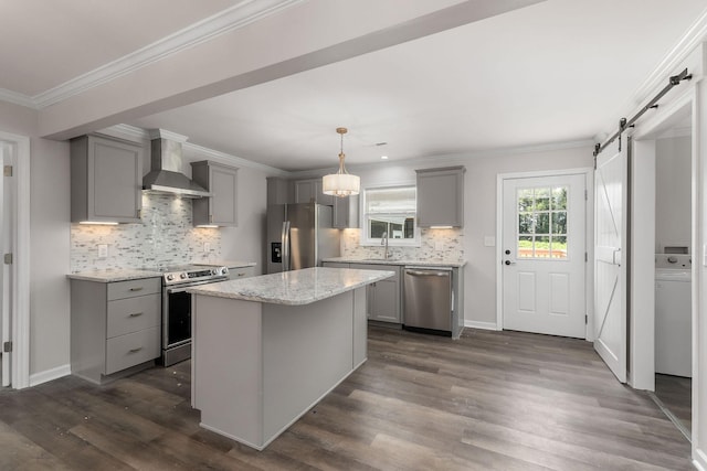 kitchen featuring wall chimney range hood, stainless steel appliances, a kitchen island, decorative light fixtures, and a barn door