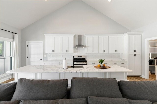 kitchen featuring stainless steel range with gas stovetop, wall chimney exhaust hood, high vaulted ceiling, an island with sink, and light wood-type flooring