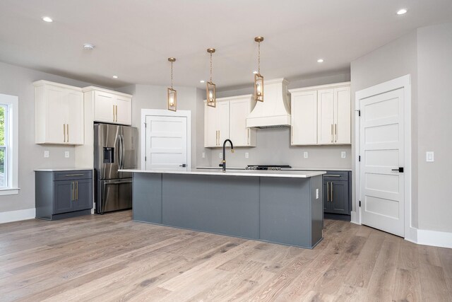 kitchen with light wood-type flooring, custom exhaust hood, a kitchen island with sink, and stainless steel refrigerator with ice dispenser