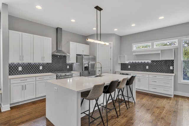 kitchen featuring dark wood-type flooring, an island with sink, stainless steel appliances, and wall chimney range hood