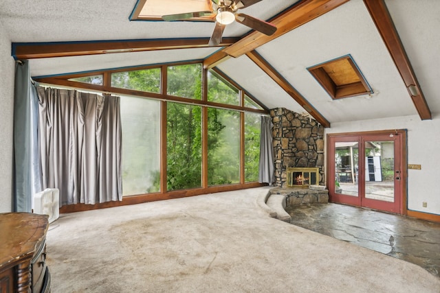 unfurnished living room with a textured ceiling, ceiling fan, lofted ceiling with beams, a stone fireplace, and carpet