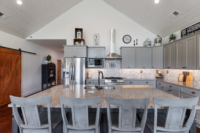 kitchen featuring stainless steel appliances, a kitchen island with sink, backsplash, a barn door, and wall chimney exhaust hood