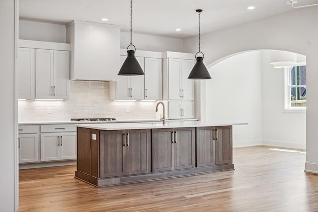 kitchen with a kitchen island with sink, custom range hood, light wood-type flooring, and decorative light fixtures