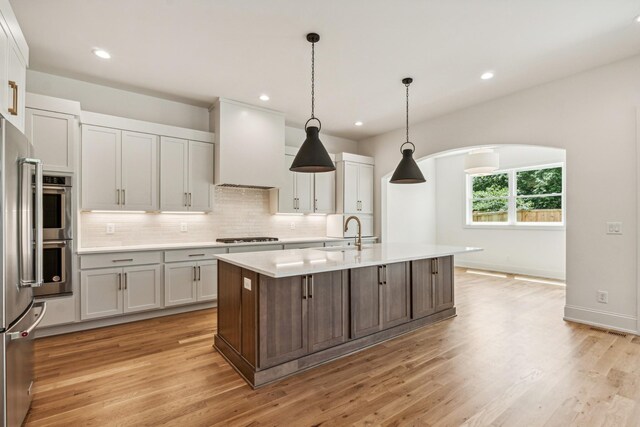 kitchen featuring pendant lighting, decorative backsplash, premium range hood, an island with sink, and light wood-type flooring