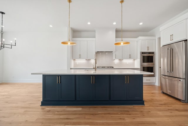 kitchen featuring a kitchen island with sink, hanging light fixtures, stainless steel appliances, white cabinets, and wall chimney exhaust hood