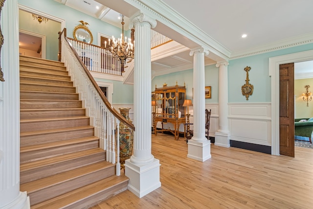 stairs with a notable chandelier, wood-type flooring, ornate columns, and ornamental molding