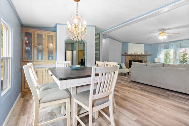 dining space with a stone fireplace, ceiling fan with notable chandelier, a textured ceiling, and light wood-type flooring