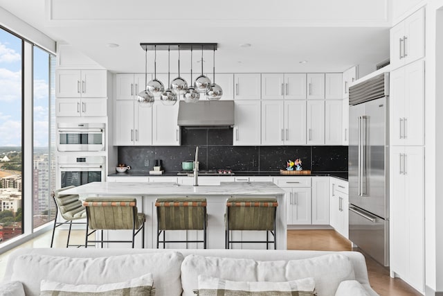 kitchen with an island with sink, light stone counters, light hardwood / wood-style flooring, wall chimney exhaust hood, and built in fridge