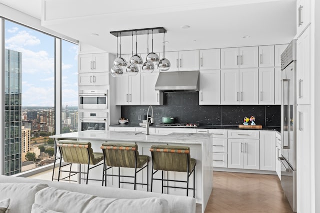 kitchen featuring wall chimney exhaust hood, white cabinets, an island with sink, and light parquet flooring