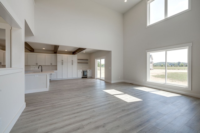 unfurnished living room with sink, light hardwood / wood-style flooring, beamed ceiling, and a high ceiling