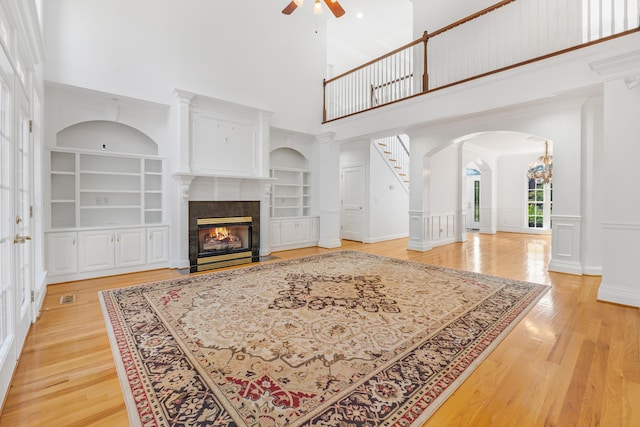 living room featuring ceiling fan with notable chandelier, a tiled fireplace, built in features, light hardwood / wood-style flooring, and a towering ceiling