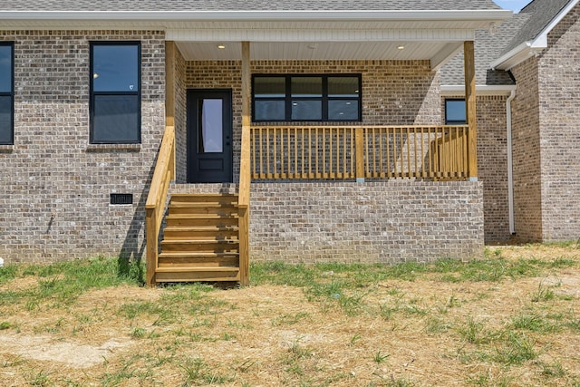 view of exterior entry with crawl space, a shingled roof, a porch, and brick siding