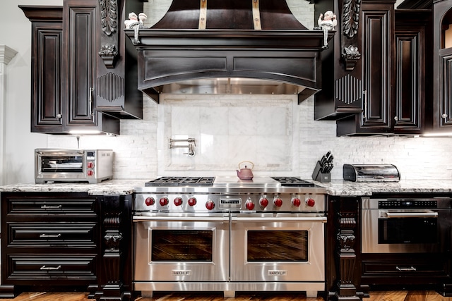 kitchen featuring appliances with stainless steel finishes, decorative backsplash, custom exhaust hood, light stone countertops, and hardwood / wood-style flooring