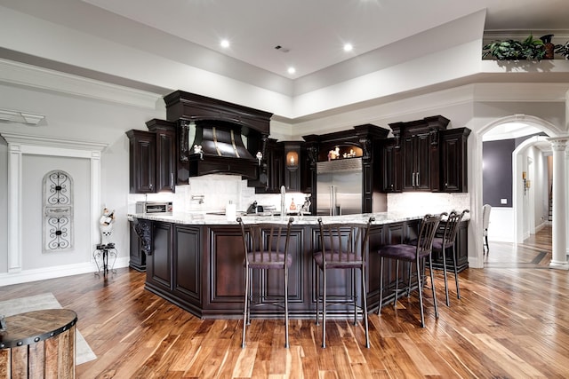 kitchen with arched walkways, premium range hood, built in refrigerator, decorative backsplash, and light stone countertops