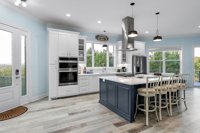 kitchen with light wood-type flooring, appliances with stainless steel finishes, island range hood, hanging light fixtures, and white cabinets