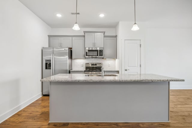 kitchen with appliances with stainless steel finishes, hanging light fixtures, light stone countertops, a kitchen island with sink, and decorative backsplash