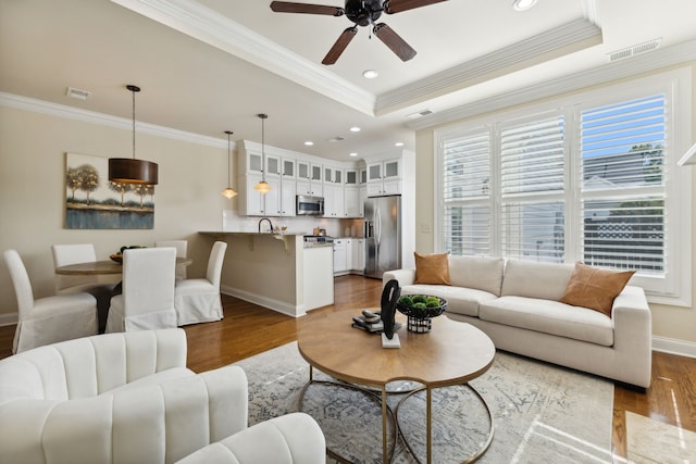 living room featuring sink, ceiling fan, light wood-type flooring, ornamental molding, and a tray ceiling