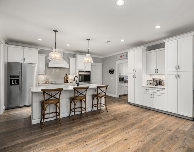 kitchen with stainless steel appliances, premium range hood, white cabinetry, a kitchen island with sink, and wood-type flooring