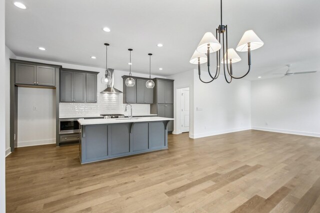 kitchen featuring a center island with sink, light wood-type flooring, pendant lighting, wall chimney range hood, and gray cabinets