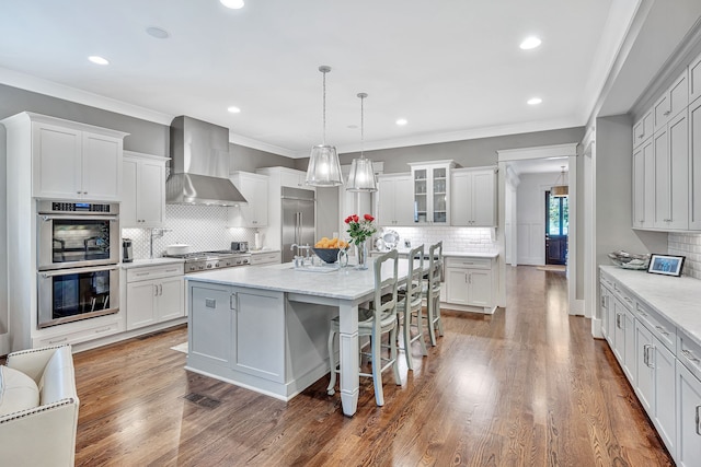 kitchen with wall chimney range hood, hardwood / wood-style floors, appliances with stainless steel finishes, and an island with sink