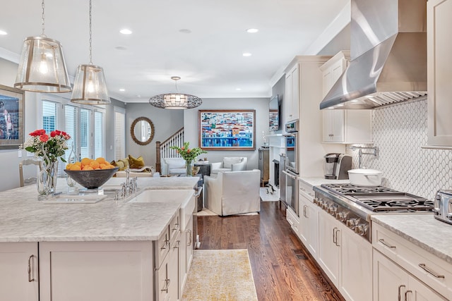 kitchen with wall chimney range hood, pendant lighting, dark wood-type flooring, and an island with sink