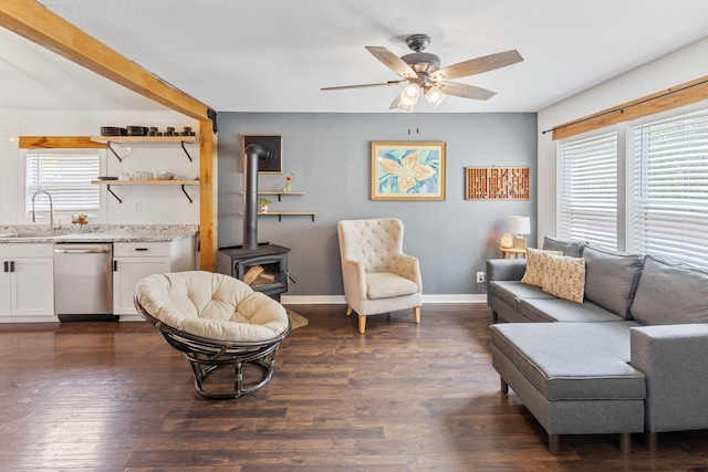 living room with ceiling fan, dark wood-type flooring, a wood stove, beam ceiling, and sink