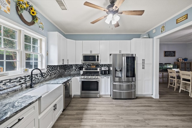 kitchen with light wood finished floors, stainless steel appliances, ornamental molding, white cabinets, and dark stone countertops