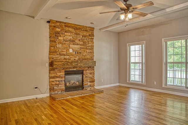 unfurnished living room featuring wood-type flooring, beam ceiling, ceiling fan, and a stone fireplace