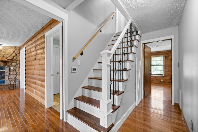 staircase featuring rustic walls, a textured ceiling, hardwood / wood-style flooring, and a stone fireplace