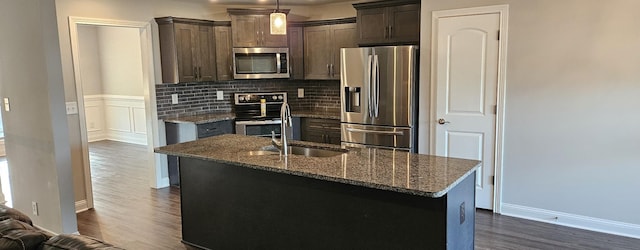 kitchen featuring dark brown cabinetry, dark stone countertops, a kitchen island with sink, stainless steel appliances, and pendant lighting