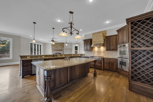 kitchen featuring dark brown cabinetry, appliances with stainless steel finishes, light stone counters, hanging light fixtures, and wall chimney range hood