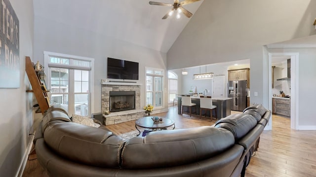 living room featuring a ceiling fan, baseboards, high vaulted ceiling, light wood-style flooring, and a fireplace