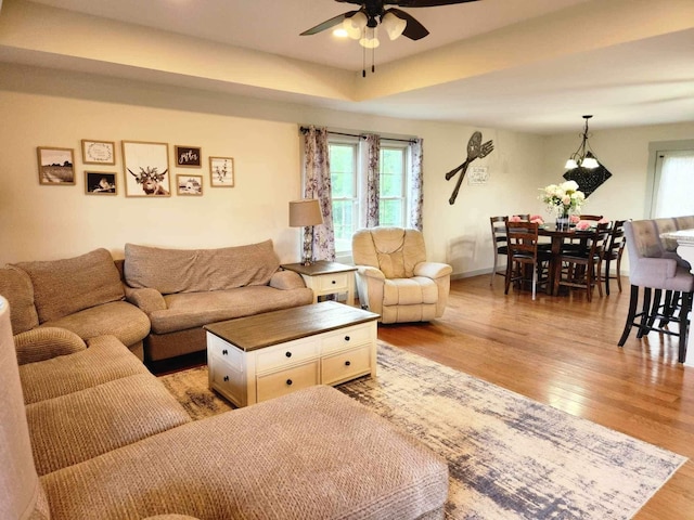 living area with ceiling fan, a tray ceiling, light wood-style flooring, and baseboards