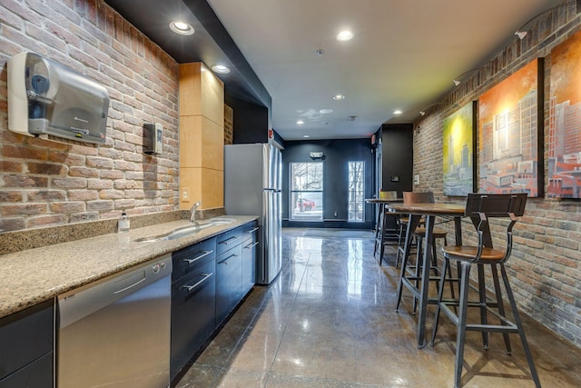 kitchen featuring brick wall, dark cabinets, light stone countertops, stainless steel appliances, and a sink